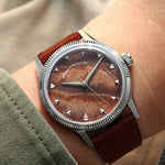 A wristwatch with a brown burlwood dial and a brown leather strap, shown worn on a person's wrist against a soft beige background.