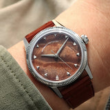 A wristwatch with a brown burlwood dial and a brown leather strap, shown worn on a person's wrist against a soft beige background.