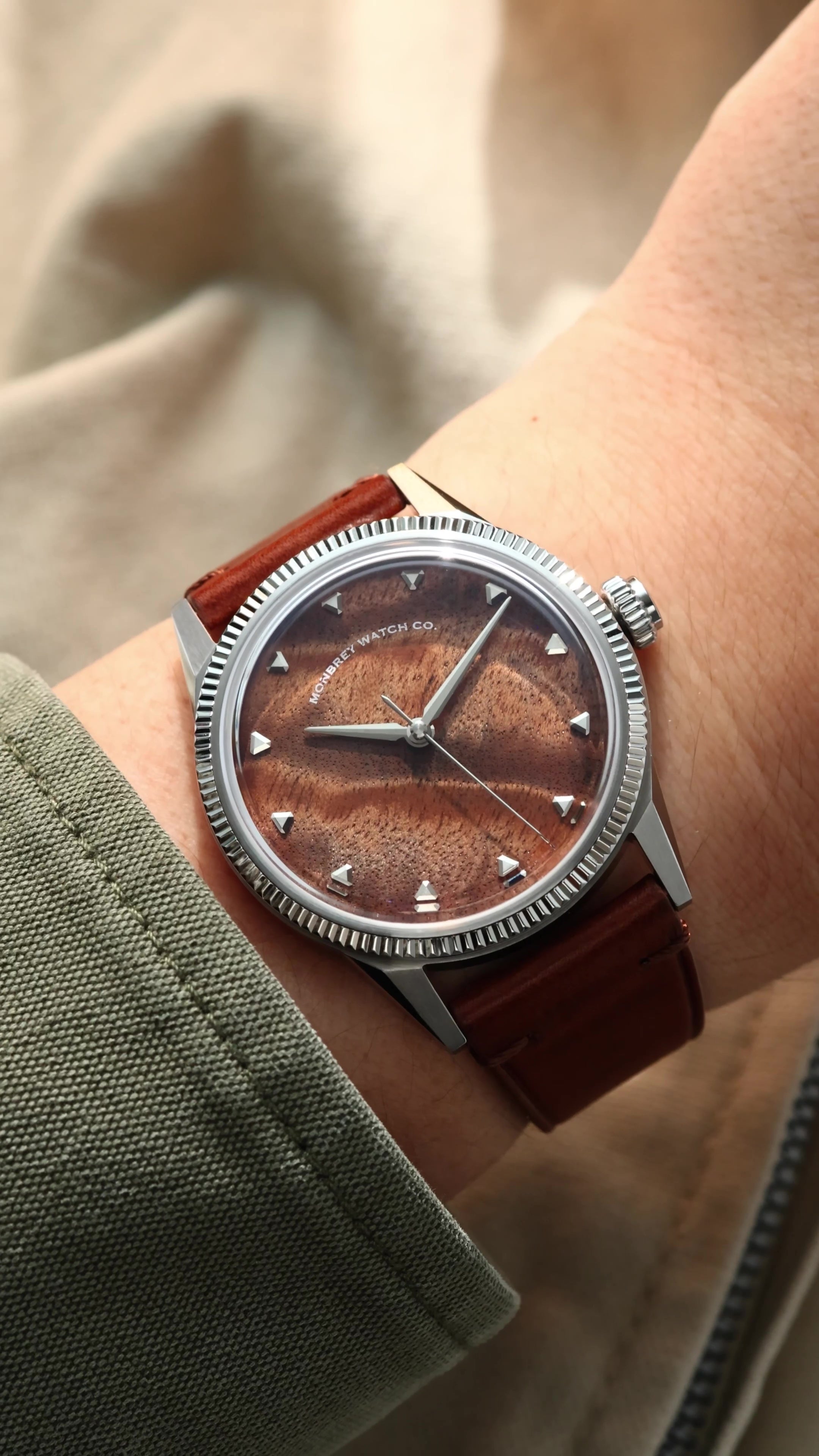 A wristwatch with a brown burlwood dial and a brown leather strap, shown worn on a person's wrist against a soft beige background.