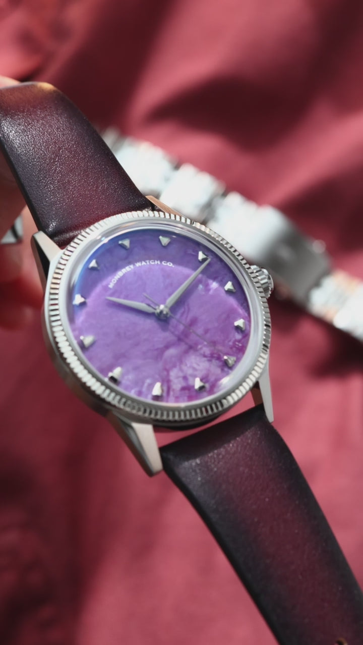 A wristwatch with a natural charoite stone dial and silver-toned case and fluted, with a purple patina leather strap, worn on a person's wrist against a dark red fabric background.