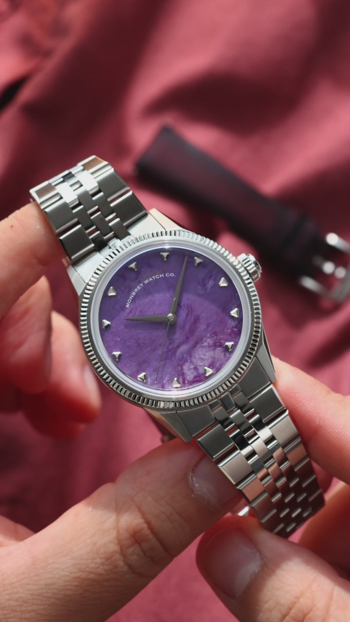 A wristwatch with a natural charoite stone dial and silver-toned case and fluted, with a silver jubilee bracelet, worn on a person's wrist against a dark red fabric background.