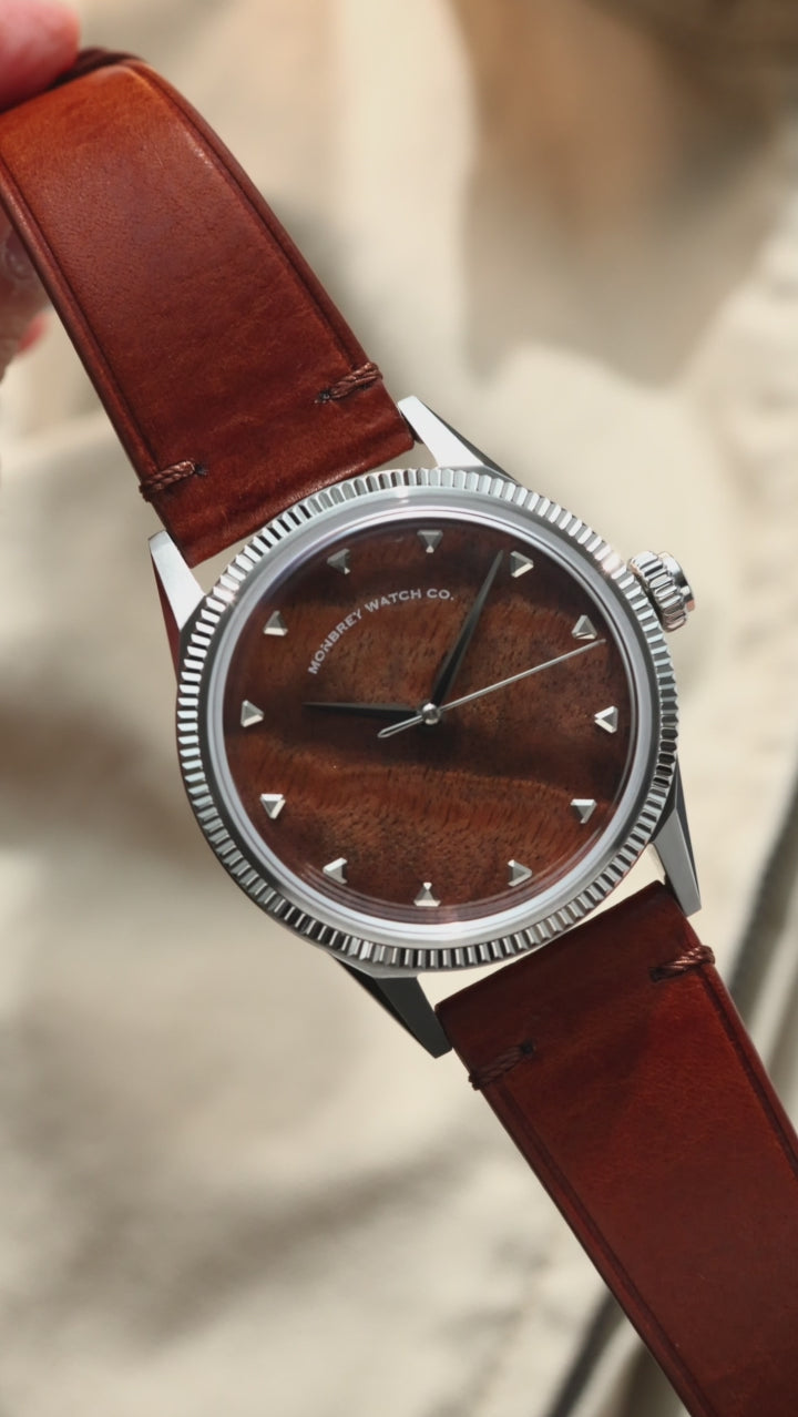 A silver-tone wristwatch with a natural burlwood dial, a fluted bezel and a brown leather strap, styled on a beige fabric surface