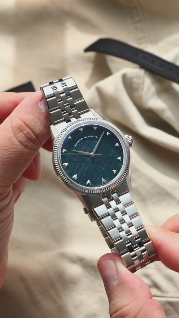 A wristwatch with a natural bloodstone dial, silver-tone case and fluted bezel, and silver jubilee bracelet, shown worn on a person's wrist against a beige fabric background.