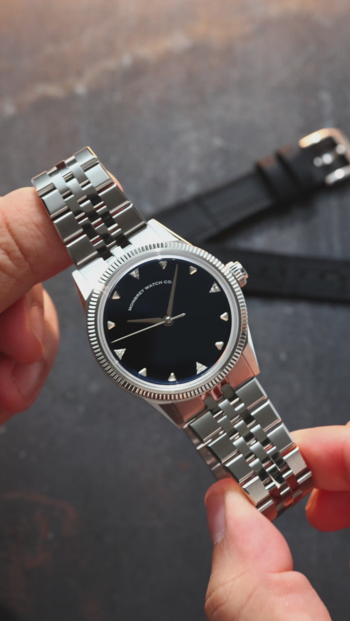 A wristwatch with a round silver-tone case and fluted bezel, a natural Onyx stone dial, and a black croco leather strap, styled on an dark textured surface background next to a silver jubilee bracelet. Lab-grown diamond on dial.