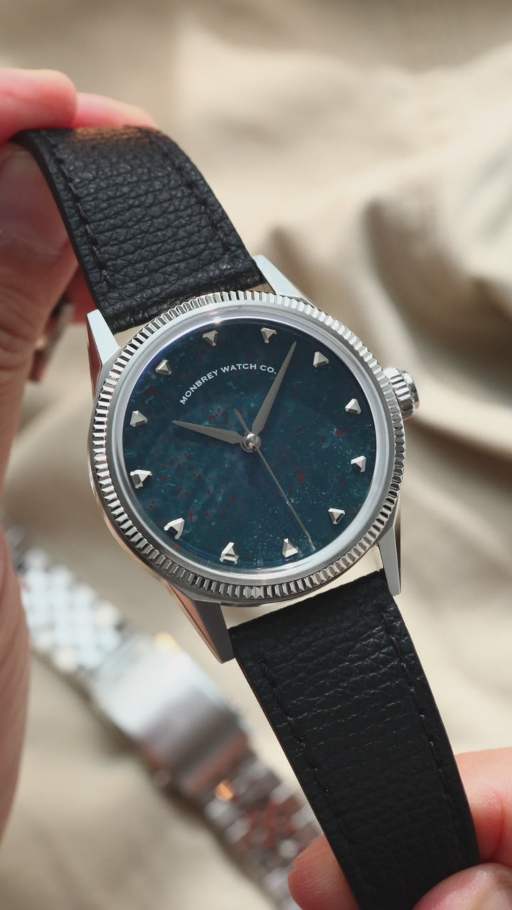 A wristwatch with a natural bloodstone dial, silver-tone case and fluted bezel, and black leather strap, shown worn on a person's wrist against a beige fabric background.