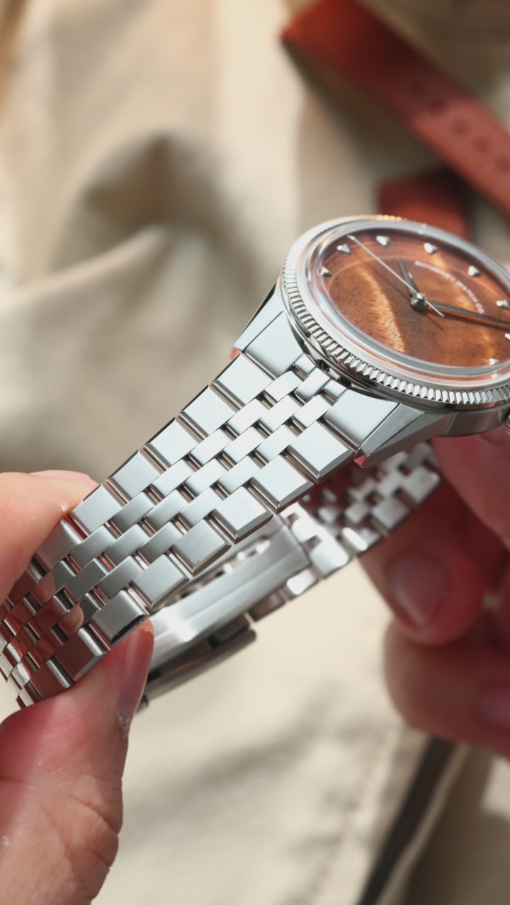 A silver-tone wristwatch with a natural burlwood dial, a fluted bezel and a jubilee bracelet, styled on a beige fabric surface